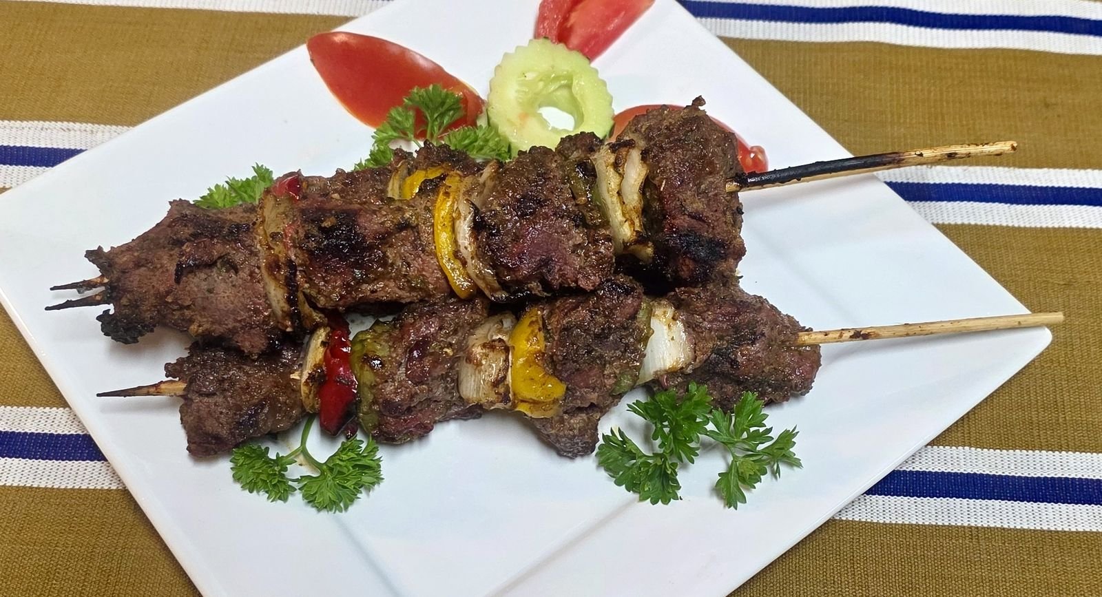 Two charcoal-grilled beef kabobs with onions and bell peppers on a white plate, garnished with tomato, cucumber and parsley