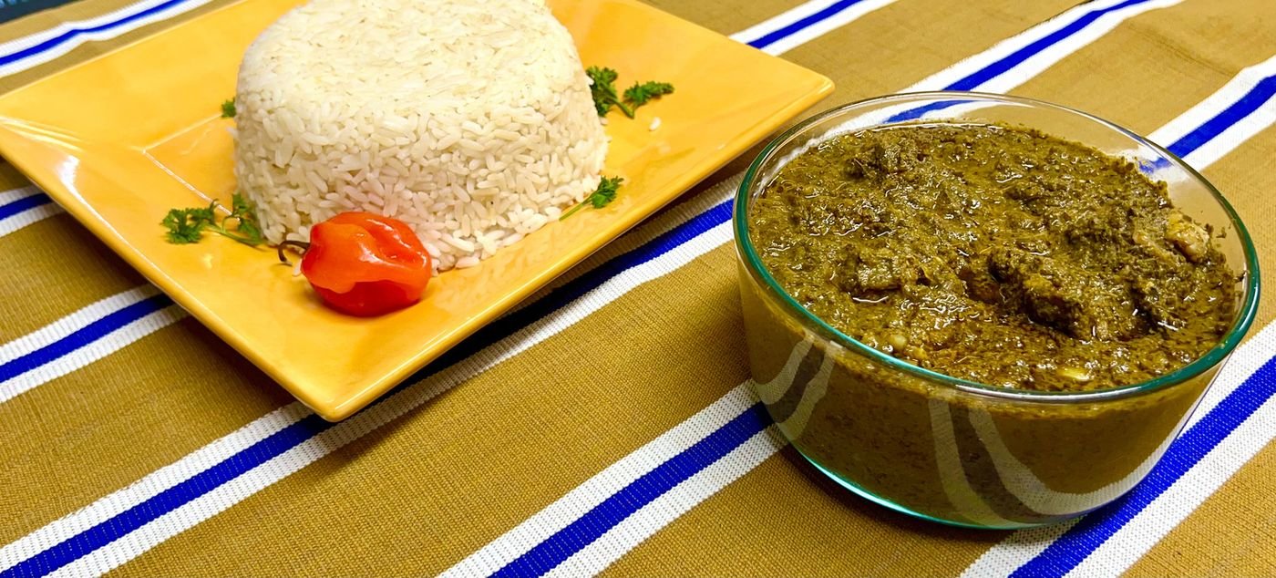 Leaf stew in a glass bowl beside a mound of white rice and a scotch bonnet pepper